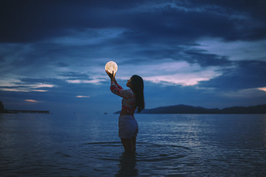 Optical Illusion Of Woman Holding Moon While Standing In Sea Against Sky At Dusk