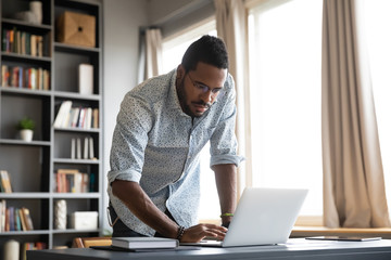 Focused african businessman standing at home office desk using laptop