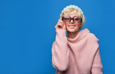 Happy girl listens to music in headphones wearing a sunglasses, knitted hat, sweater over blue background
