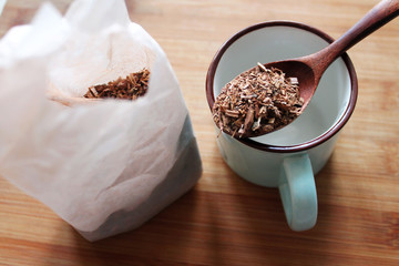 Dried ground oak tree bark in a wooden spoon lying on a blue cup. Paper packet half full of oak bark on a woden board.