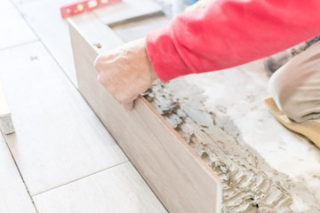 Man installing rectangular shaped floor tiles in kitchen. Applying adhesive before installation and verifying afterwards