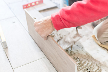Man installing rectangular shaped floor tiles in kitchen. Applying adhesive before installation and verifying afterwards
