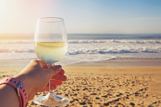 Female hand holding glass of white wine with sea on background.