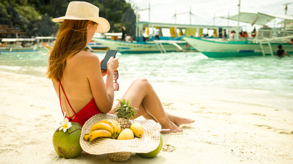 Young redhead freelancer female in bikini and hat working remotely with her phone - Banul beach, Coron, Palawan, Philippines