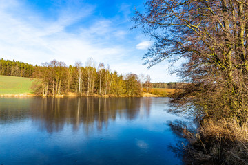 Spring lake in czech countryside. Blue sky, green field and frozen water.