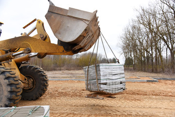 Excavator bucket lifts the pallet with road tiles, road construction.