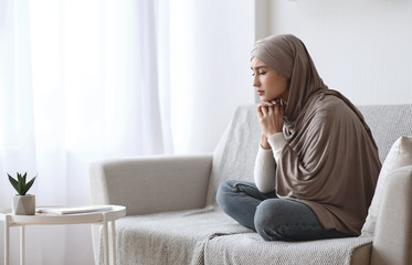 Depressed muslim woman in hijab sitting upset on sofa at home