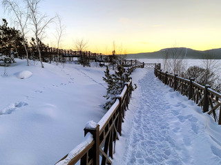Snowy new wooden bridge on a lake with a beautiful evergreen pine forest and mountains on horizon. Sunset in winter time. 