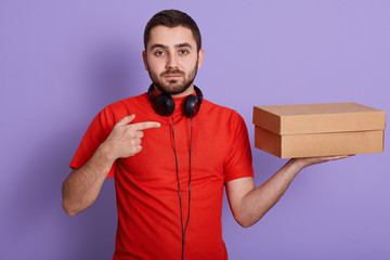 Portrait of serious courier wearing red t shirt holding box and looking at camera, pointing at...