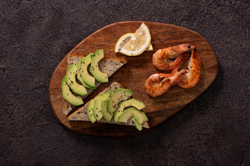 Wholegrain toast bread slices with guacamole, fried shrimp and fried bacon pieces on wooden board Selective Focus, Focus on the front of the shrimp on the first bread