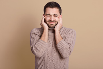 Young Caucasian man wearing warm knitted sweater standing isolated over beige background, bearded man covering ears with fingers with annoyed expression and squinted eyes, hearing noise of loud music.