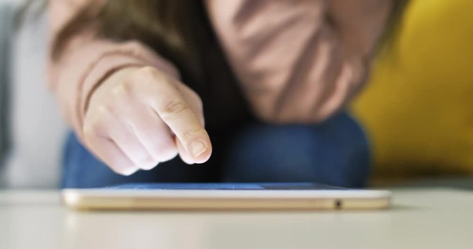 Young woman using rose gold tablet and seat on couch or sofa in jeans and pastel pink sweater. Woman using tablet on table for social network or shopping online.
