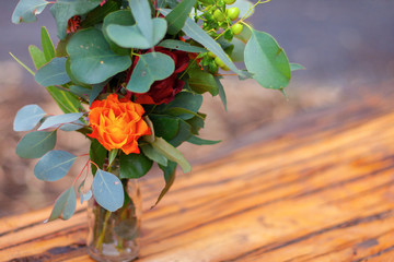 A green bouquet of beautiful bright flowers in a glass bottle standing on the tree trunk in the middle of the forest. Focus on a bright orange flower. Closeup.