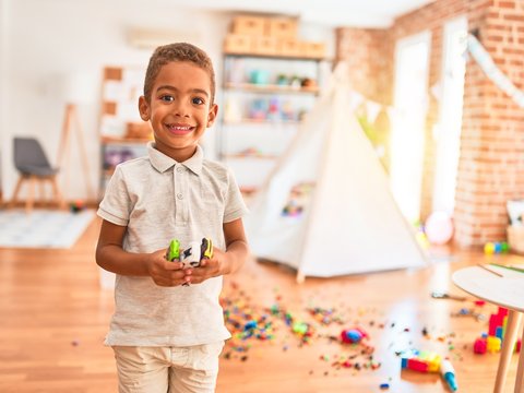 Beautiful african american toddler holding cars toy smiling around lots of toys at kindergarten