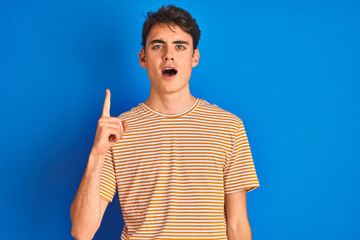 Teenager boy wearing casual t-shirt standing over blue isolated background pointing finger up with...