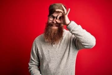 Handsome Irish redhead man with beard wearing casual sweater and glasses over red background doing ok gesture with hand smiling, eye looking through fingers with happy face.