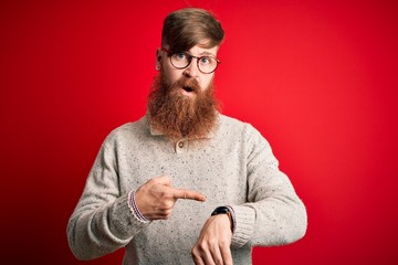 Handsome Irish redhead man with beard wearing casual sweater and glasses over red background In...