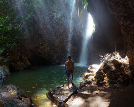  Backside of man traveler stand under the ray of light in the cave. Form Bali. Travel concept. The Gitgit waterfall on the island of Bali. Waterfall in the tropical forest.