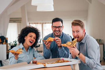 Group portrait of friends eating pizza indoors, smiling at camera.