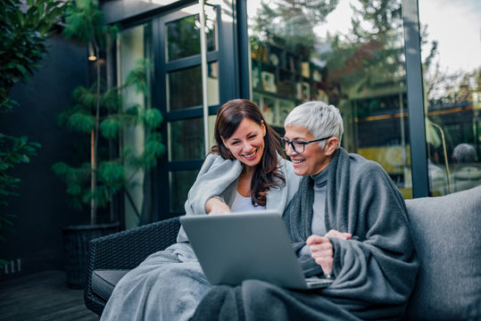 Happy beautiful older mother and adult daughter using laptop while sitting on wooden terrace, portrait.