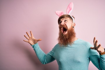 Hipster Irish man with beard wearing easter rabbit ears over isolated pink background crazy and mad...