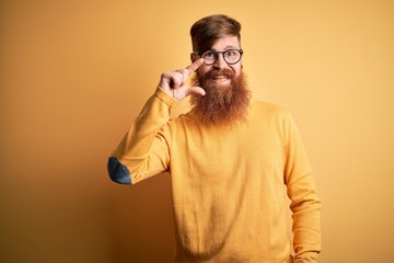 Handsome Irish redhead man with beard wearing glasses over yellow isolated background smiling and confident gesturing with hand doing small size sign with fingers looking and the camera. Measure