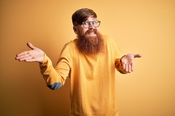 Handsome Irish redhead man with beard wearing glasses over yellow isolated background clueless and...