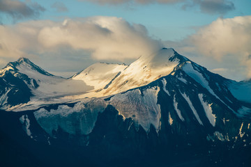 Atmospheric alpine landscape with big snowy mountains among low clouds in golden hour. Wonderful highland scenery with massive glacier on giant mountain range in sunrise. Shiny snow on big rockies.