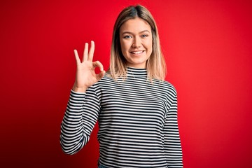 Young beautiful blonde woman wearing casual striped sweater over red isolated background smiling positive doing ok sign with hand and fingers. Successful expression.