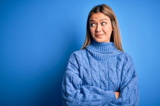 Young beautiful blonde woman wearing winter wool sweater over blue isolated background smiling looking to the side and staring away thinking.