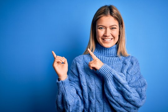 Young beautiful blonde woman wearing winter wool sweater over blue isolated background smiling and looking at the camera pointing with two hands and fingers to the side.