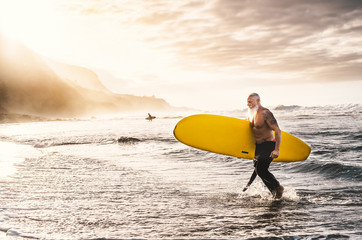 Happy fit senior surfing on sunset time - Sporty mature man having fun training with surfboard in ocean - Elderly healthy people lifestyle and extreme sport concept