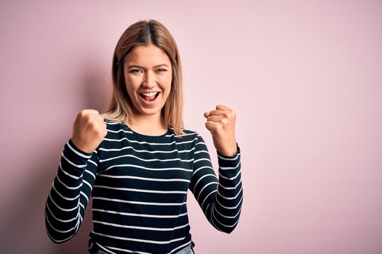 Young beautiful blonde woman wearing casual striped sweater over pink isolated background celebrating surprised and amazed for success with arms raised and open eyes. Winner concept.