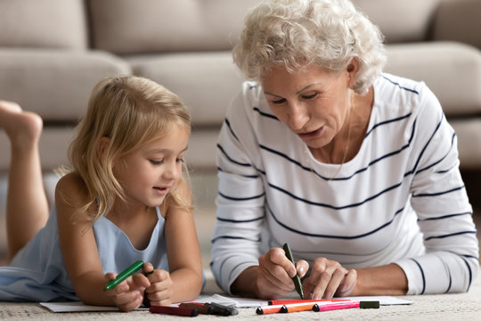 Old senior woman teaching small granddaughter coloring images.