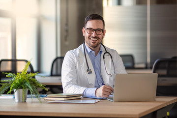 Happy male medical doctor portrait in hospital. Portrait of a male doctor with laptop sitting at desk in medical office. Portrait of a happy young doctor at medical office desk.