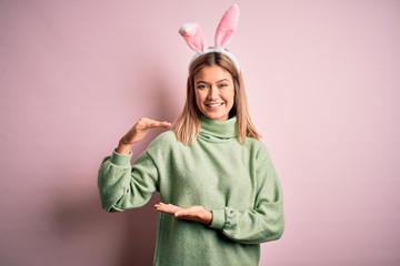 Young beautiful woman wearing easter rabbit ears standing over isolated pink background gesturing with hands showing big and large size sign, measure symbol. Smiling looking at the camera. Measuring