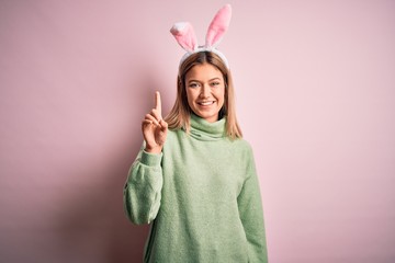 Young beautiful woman wearing easter rabbit ears standing over isolated pink background showing and pointing up with finger number one while smiling confident and happy.