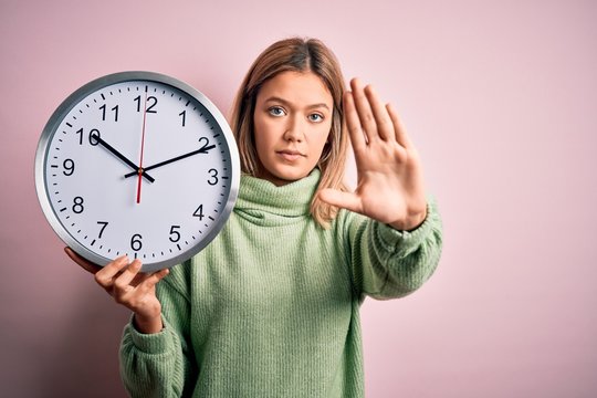 Young beautiful woman holding clock standing over isolated pink background with open hand doing stop sign with serious and confident expression, defense gesture