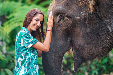 A happy Caucasian woman in a bright shirt poses next to an elephant with her hand on its trunk....