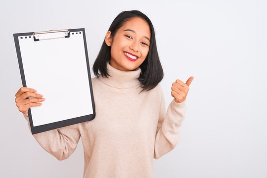 Young chinese inspector woman holding clipboard standing over isolated white background happy with big smile doing ok sign, thumb up with fingers, excellent sign