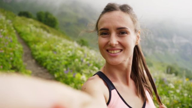 Sport woman relaxing on mountain top above the clouds. POV girl standing on trail through flowering alpine meadow, taking selfie on smartphone, looking at camera while enjoying picturesque view 