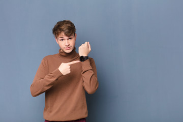 young blonde man looking impatient and angry, pointing at watch, asking for punctuality, wants to...