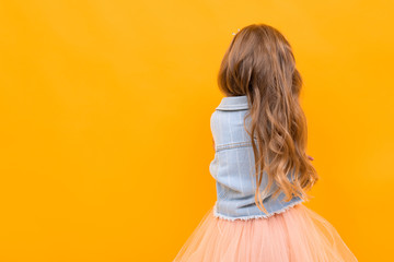 Fototapeta na wymiar girl with beautiful blonde hair in a denim jacket and a pale pink skirt stands with her back on a yellow background with copy space