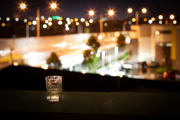 A glass of spirit on a balcony (terrace, veranda) overlooking the city highway at night with bright lights. Leisure and loneliness in the big city.