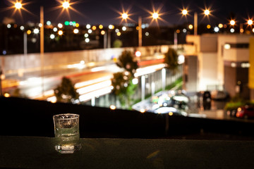 A glass of spirit on a balcony (terrace, veranda) overlooking the city highway at night with bright lights. Leisure and loneliness in the big city.