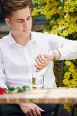 Good-looking young man wearing white shirt sitting at cafe table with glass of water and rose on it waiting for his girlfriend