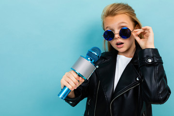 Nice girl in black jacket, blue sunglasses sings songs with blue microphone isolated on blue background