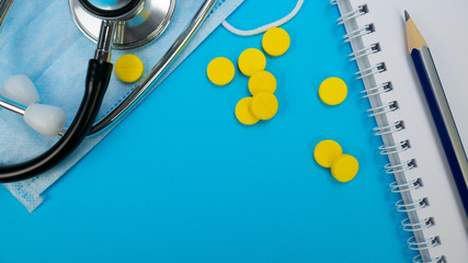 Medical face mask, phonendoscope, yellow tablets or pills on blue background. Pharmaceutical medicament. Novel coronavirus denoted 2019-nCoV concept. Search vaccine to treat disease with vaccination