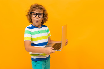 Little boy with curly hair in colourful t-shirt and shorts holds a laptop isolated on yellow...