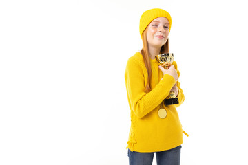 Happy teenager girl with red hair, yellow hoody and trousers holds a gold cup isolated on white background